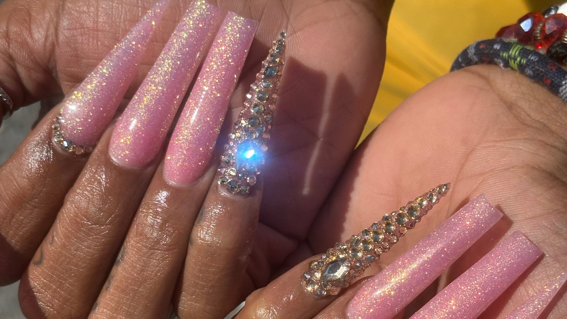 A close-up image of acrylic nails with a pink color and gold glitter, showcasing a butterfly pattern and curved shapes.